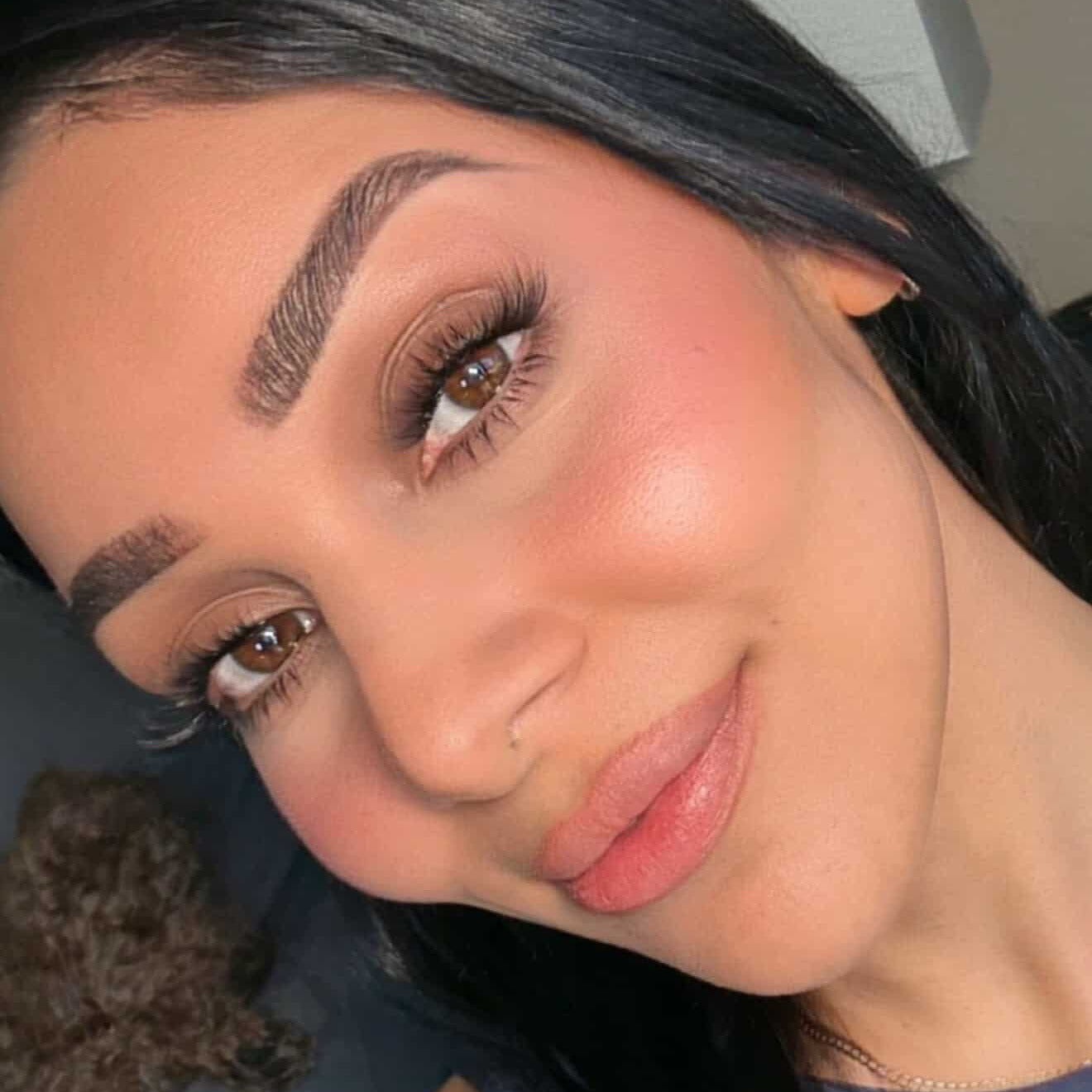 A high-angle close-up portrait of a model showcasing a completed iLash Mafia BUSSIN DIY lash extensions application. The wispy, layered volume lashes feature clean separation and soft, feathered tips that beautifully enhance her brown eyes, complemented by detailed laminated brows, luminous pink blush, and a satin-finish nude lip
