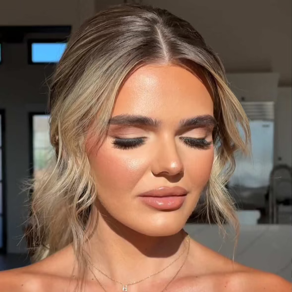 A professional beauty portrait of a model with her eyes closed, showcasing the iLash Mafia BUSSIN DIY lash extensions. The look features dense, wispy volume lashes that taper elegantly across the lash line, paired with a sun-kissed makeup look, a soft updo with blonde face-framing pieces, and delicate gold necklaces under warm, direct studio lighting.